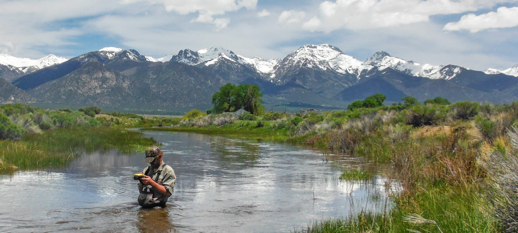 Fly fisherman hip-deep in a river. Mountain snow-capped peaks landscape.