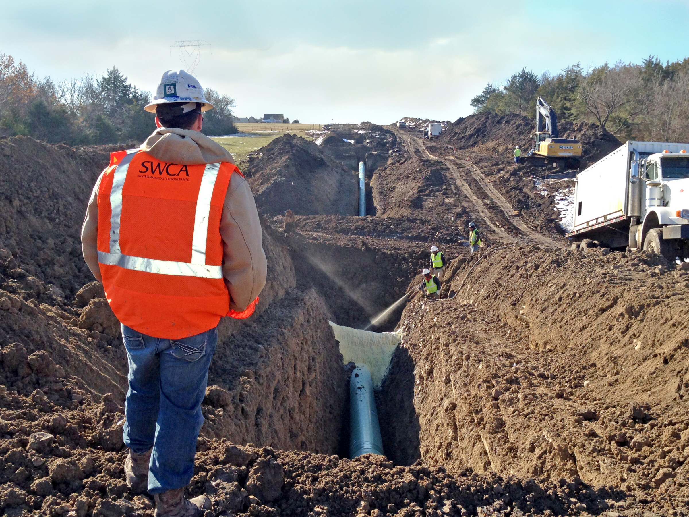 Staff member in an orange safety vest standing in front of a trench with a pipe at the bottom. 