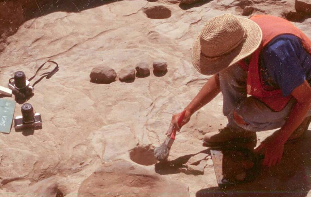 Archaeologist crouches to clear a dig site with a brush