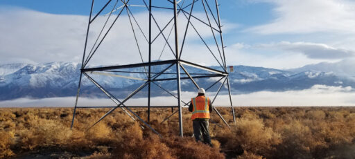 A worker in a hard hat stands at the foot of an electrical transmission tower.