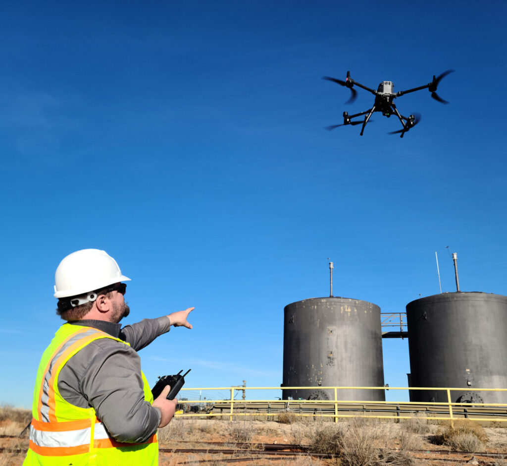 A man in a hard hat and safety vest is pointing in the air towards the drone he is flying. Cylindrical tanks in the background.