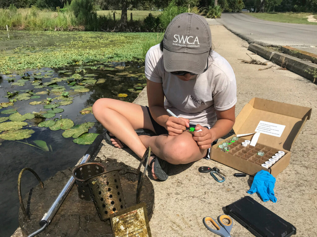 Person with an SWCA hat sitting on the ground near a pond, measuring a water sample.