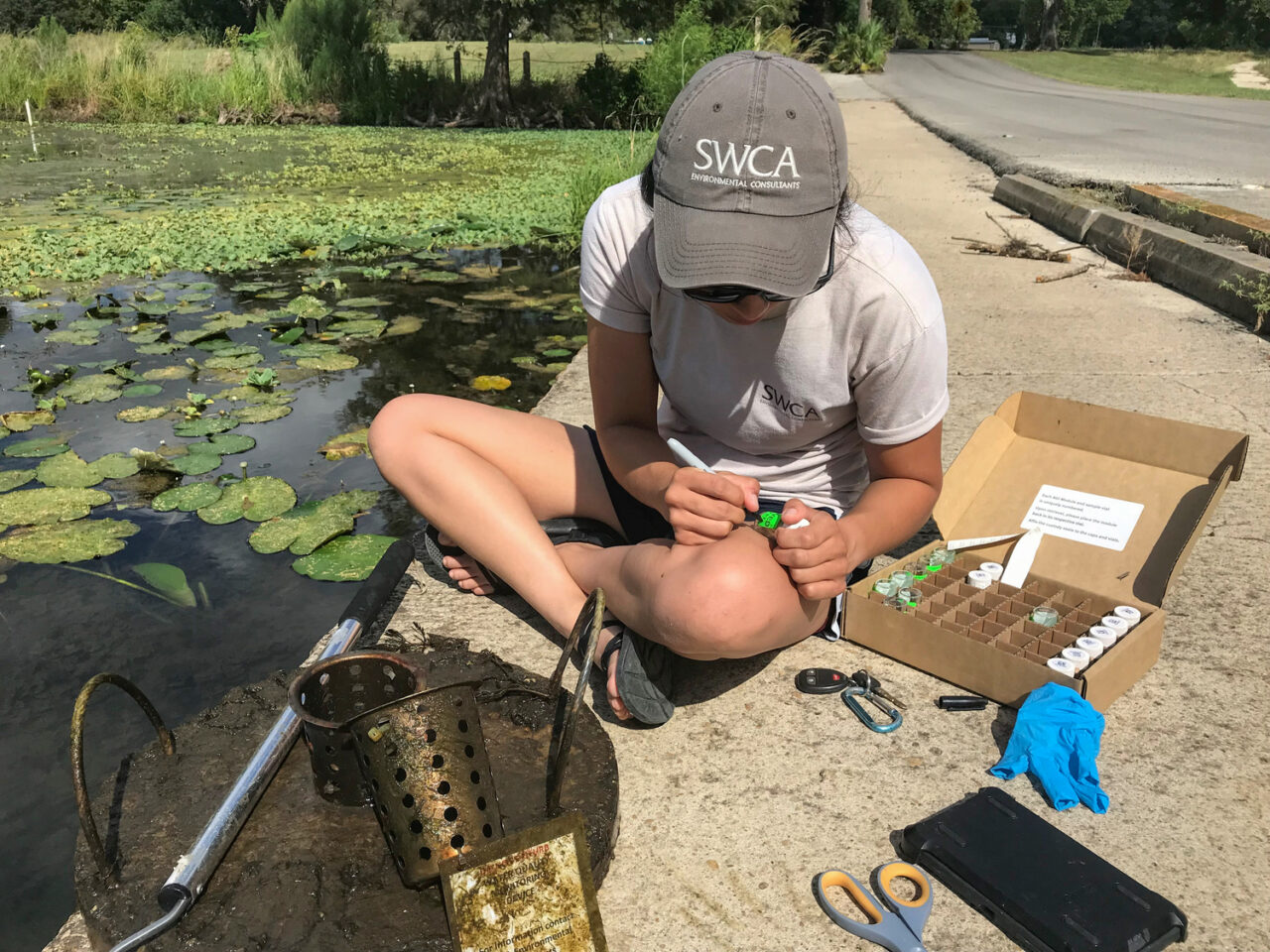 Person with an SWCA hat sitting on the ground near a pond, measuring a water sample.