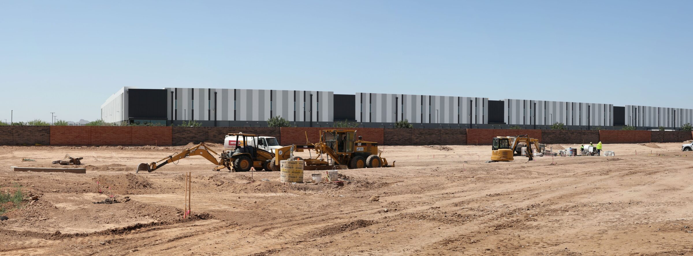 View from within the southeast corner parking lot of Goodyear Police Department looking southwest with bulldozers and workers ona dirt lot. A brick wall is also seen in the background with a large building behind it.
