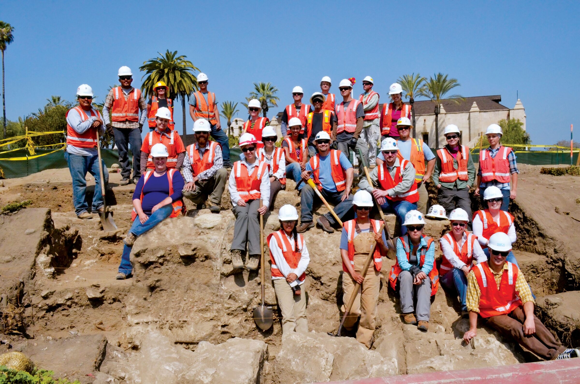 Group employee photo of over twenty field workers on an excavation site with palm trees in background.