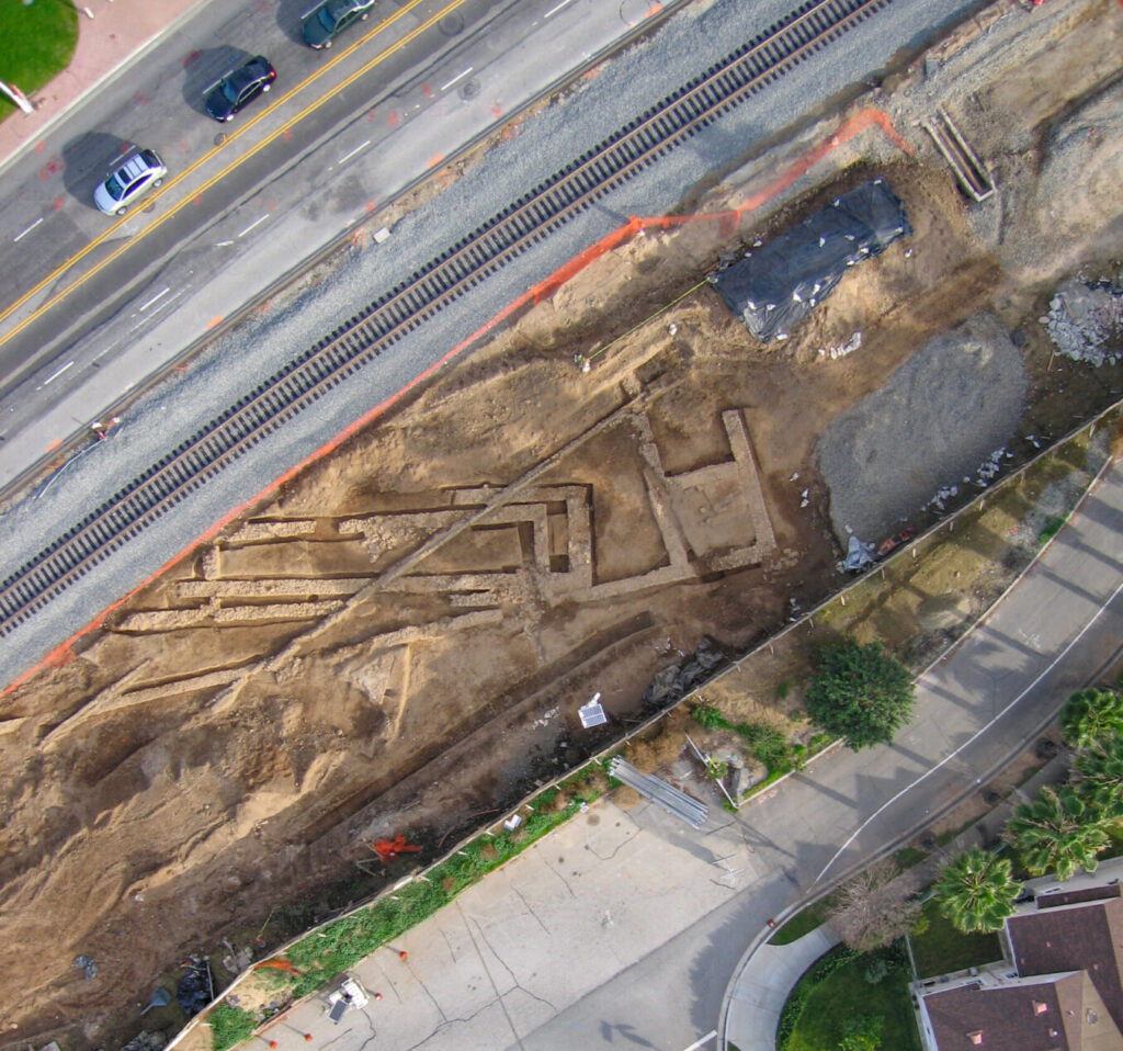 Aerial view of a long trench with exposed pipes, dug between a road, railroad, and a sidewalk.
