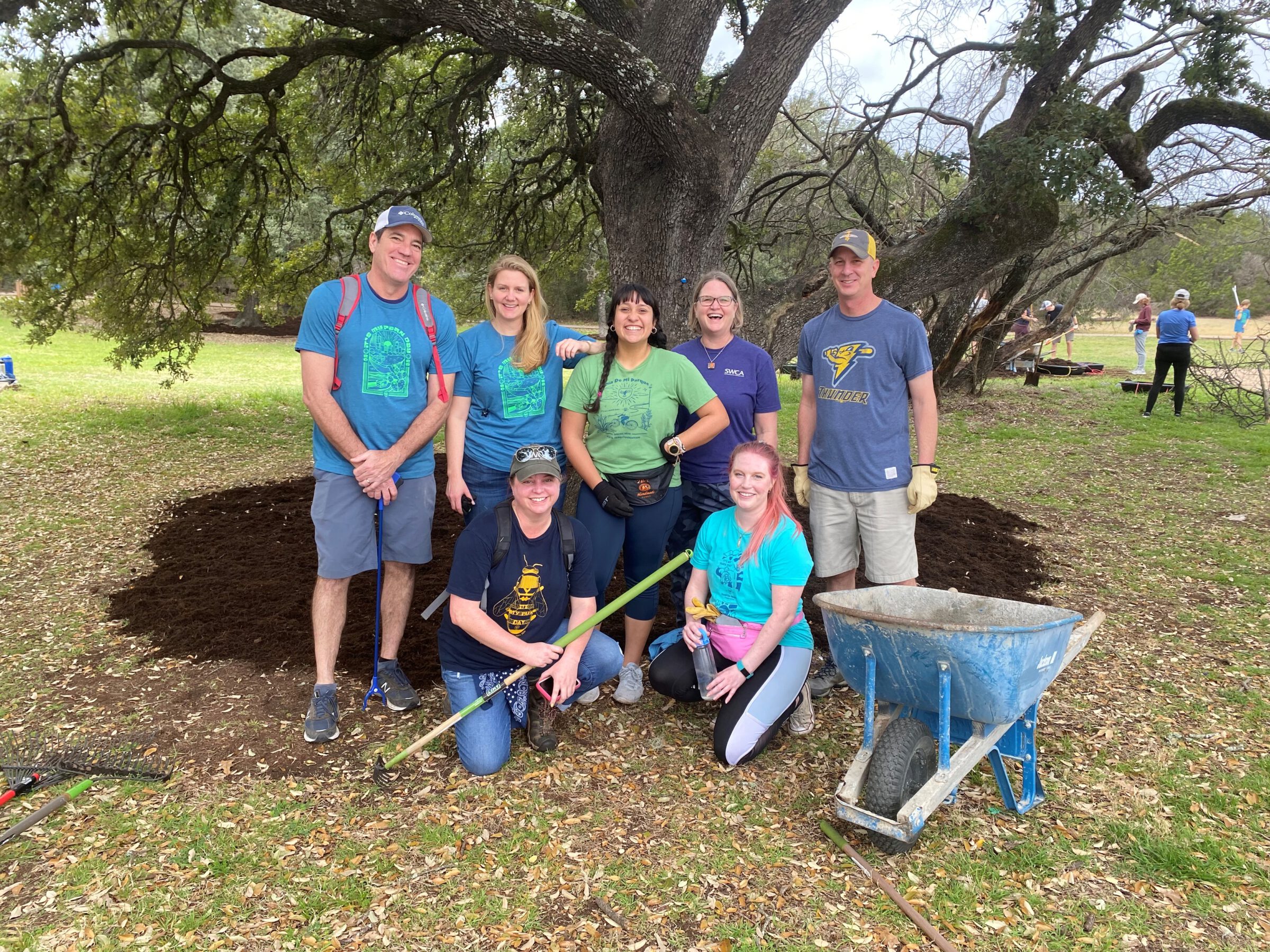 Seven people pose near a tree with new mulch, and a wheelbarrow.