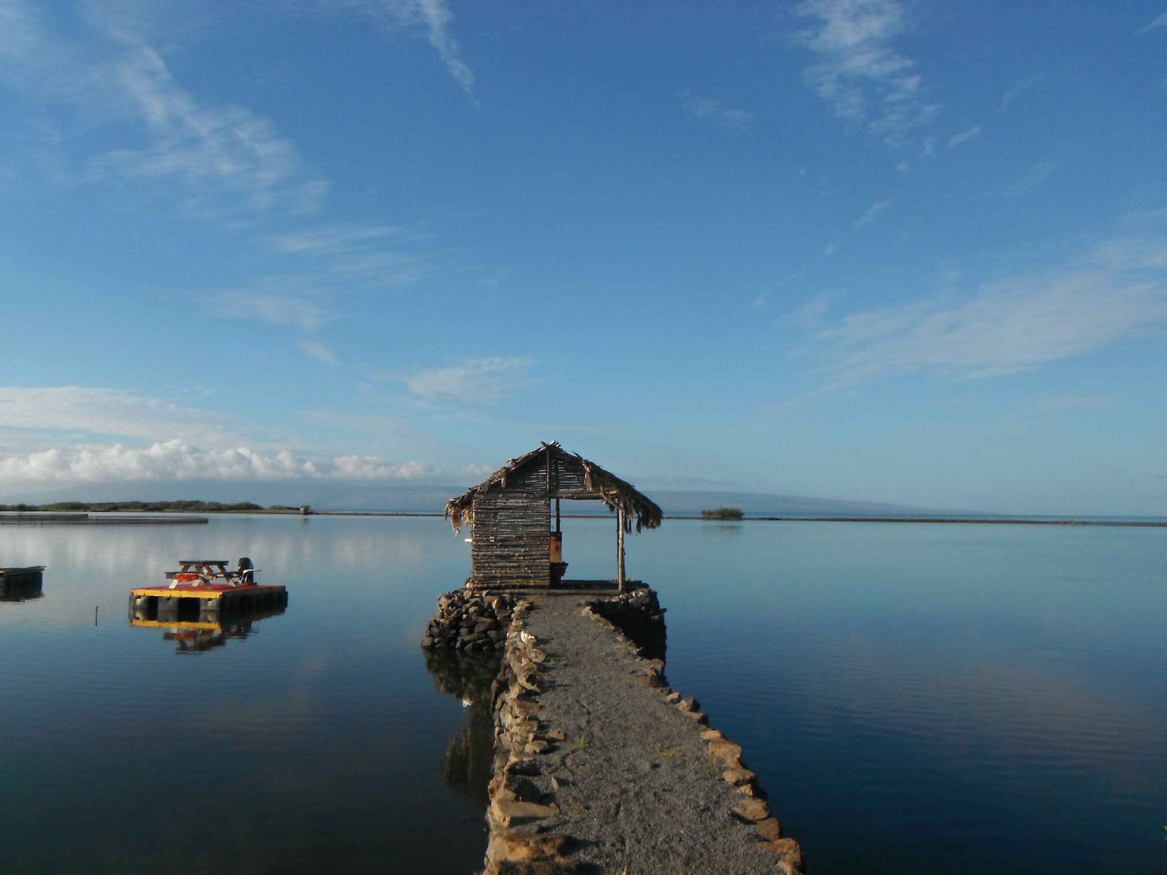 A hut on top of a small pier made of rocks. A small table floats on a concrete slab next to it.A large body of water is surrounding the area.