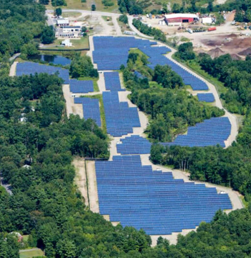 Aerial view of a large solar panel array with many rows, set in a forested area on the edge of a town.