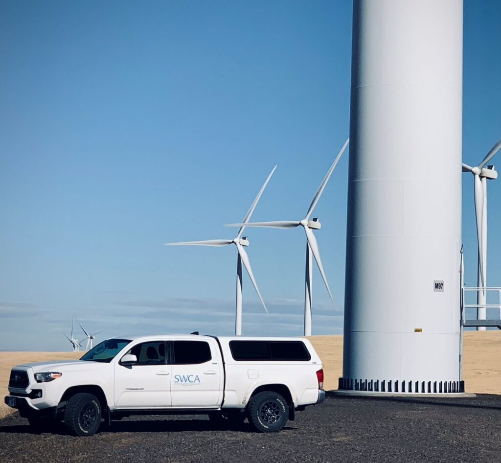An SWCA SUV parked at the base of a wind turbine, with five other turbines behind it in the distance.