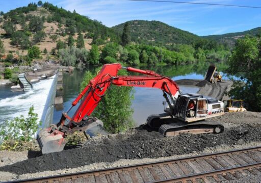Backhoe digging dirt near a body of water and train tracks.