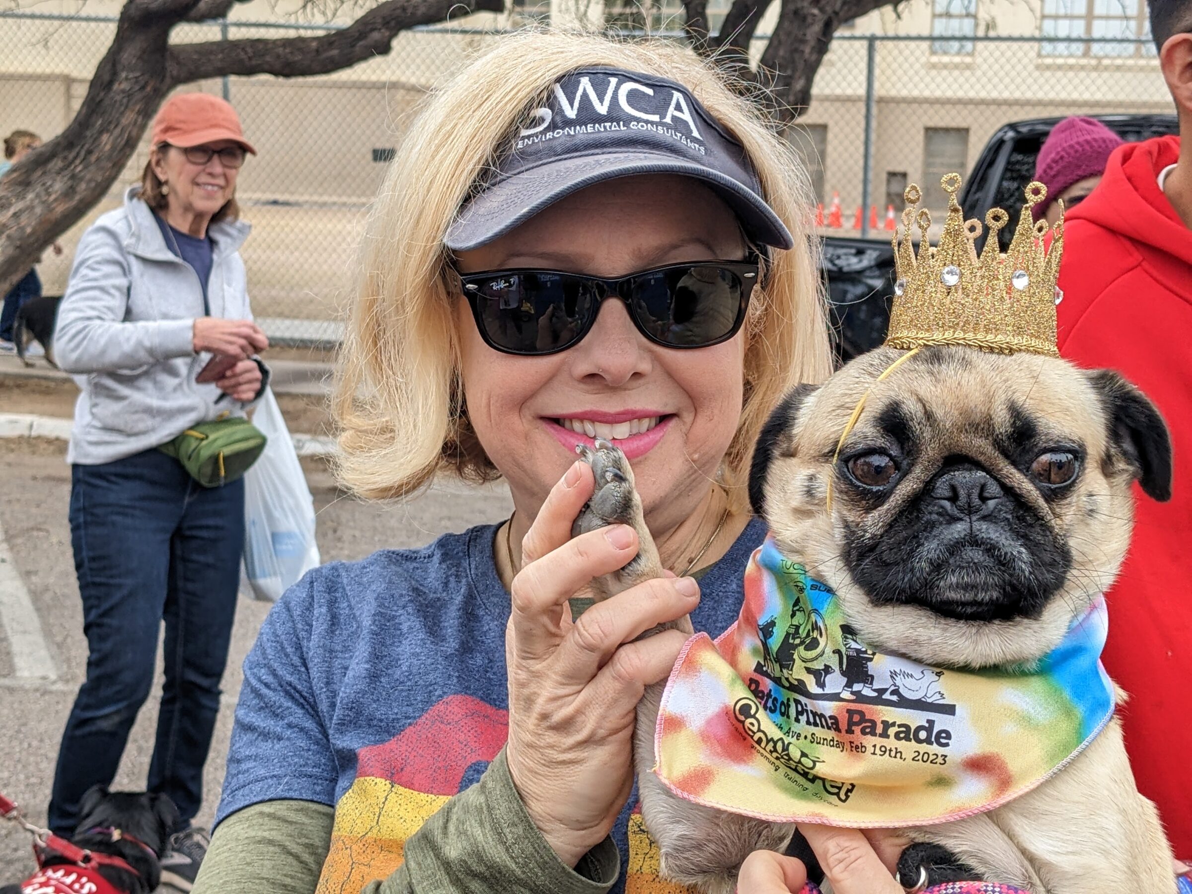 A woman wearing SWCA visor holds a pug dog wearing a crown and bandana.