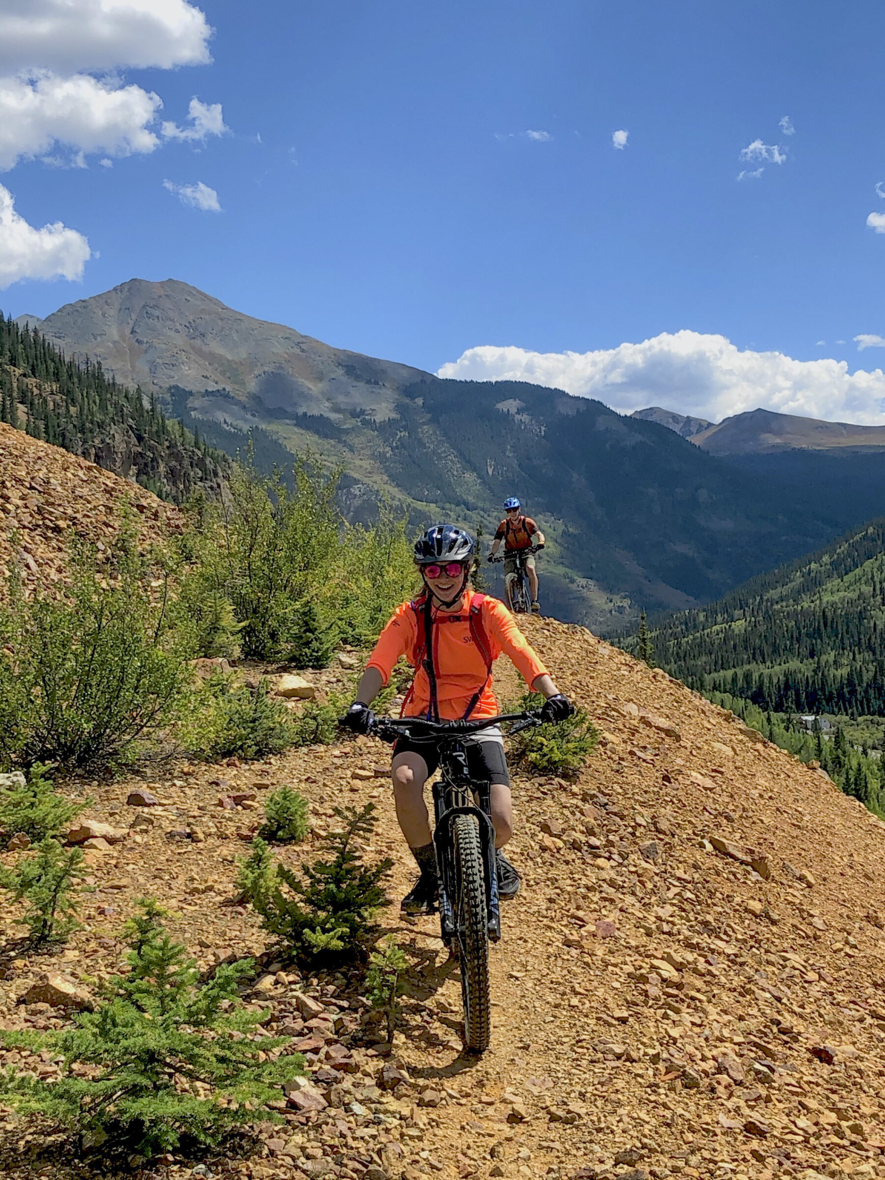 Two mountain bikers approach the camera. They ride on a rocky path on a mountainside.