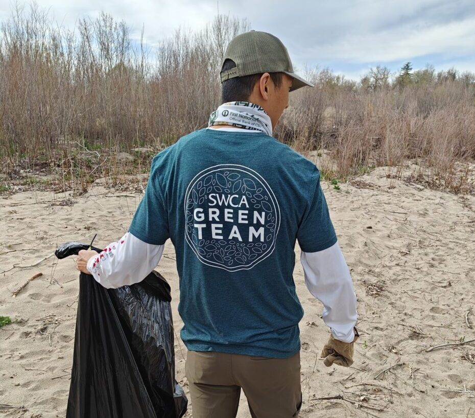 Man on the beach holding a trash can. Tee shirt says "SWCA Green Team".
