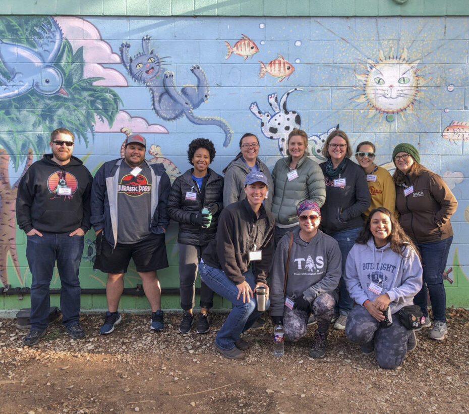 Eleven people pose for a group photo outdoors in front of a wall mural.