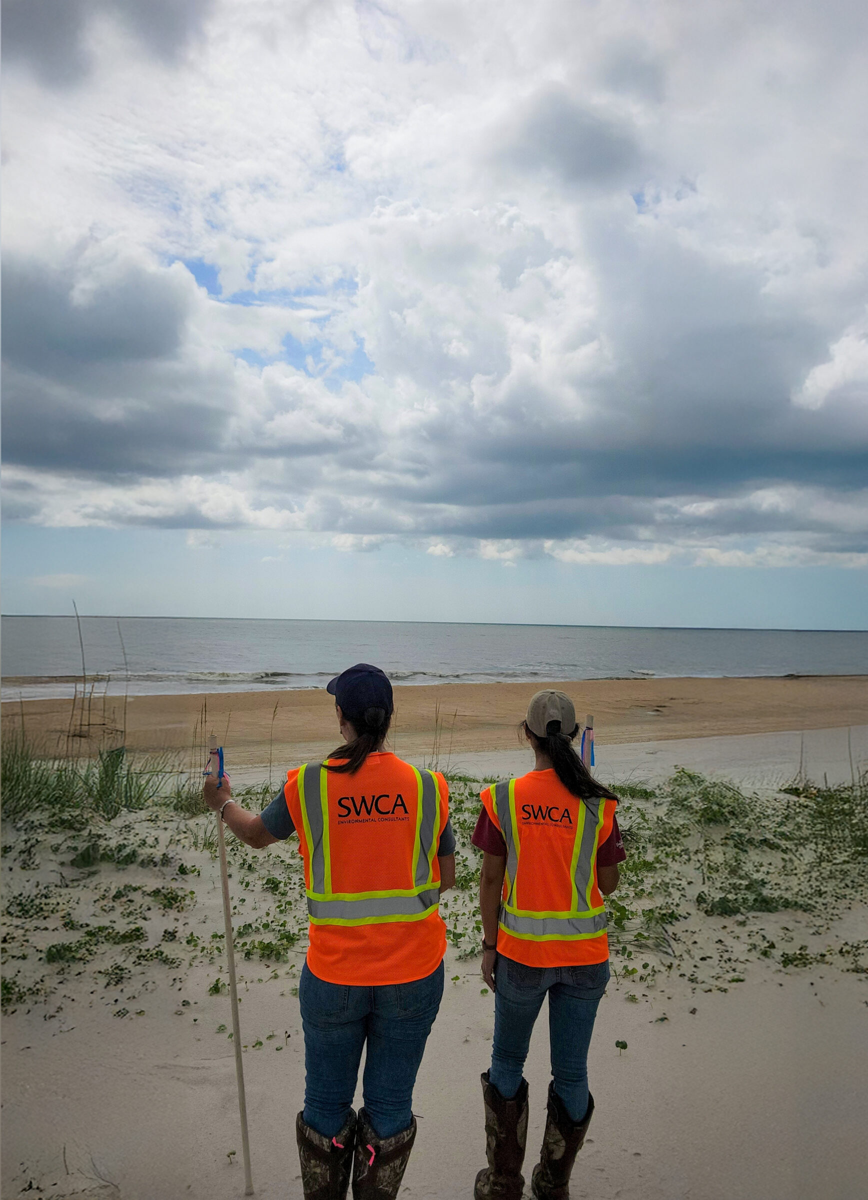 two women in SWCA safety vests survey the weather and ocean beach ahead of them.