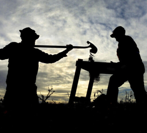 One person holding a shovel and pouring dirt through a sifter that another person is holding. The two look like shadows with the sun setting behind them.