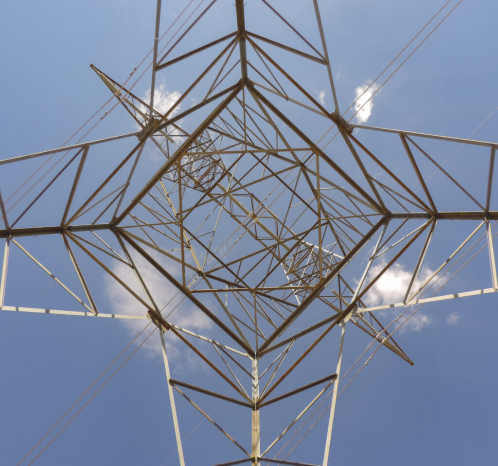 View from directly under a transmission tower, looking up. Iron scaffolding forms geometric patterns.