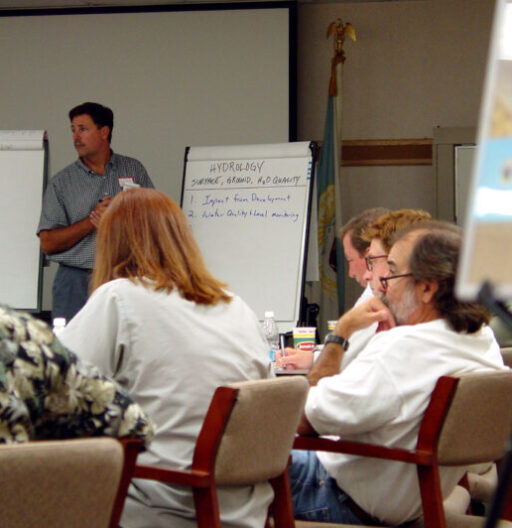 Rear view of meeting attendants seated at a table, and one man facing them is taking notes on a paper flipchart.