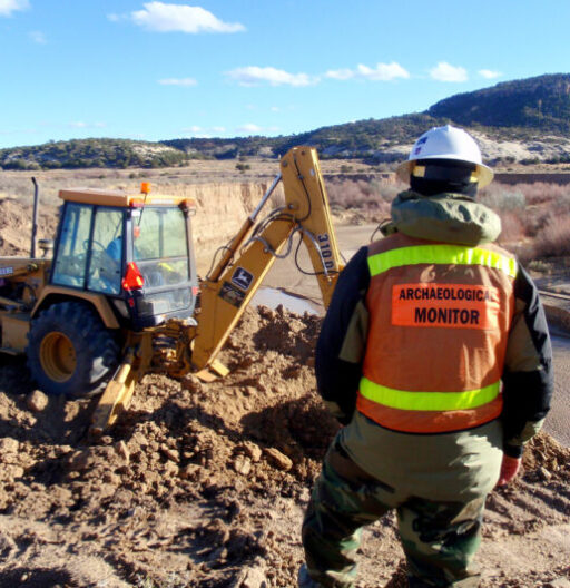 Man in safety vest labelled Archaeological Monitor at a dig site near a backhoe.
