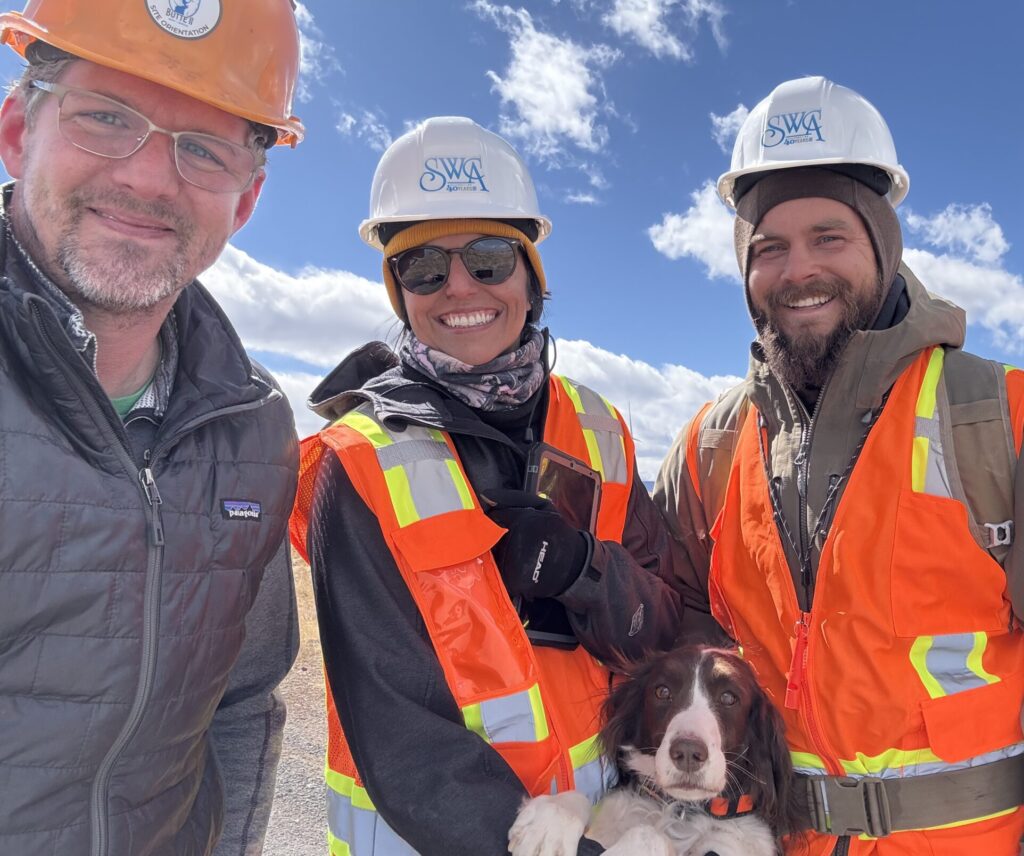 Three people outdoors in hard hats pose for a group selfie with a dog.
