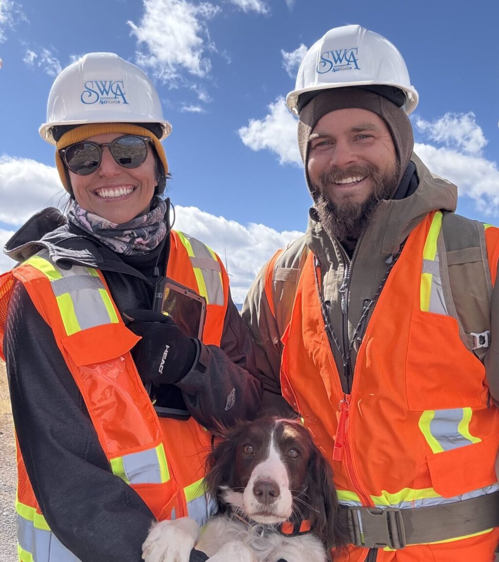 Two SWCA workers in orange safety vest and hard hat smile with a dog between them.