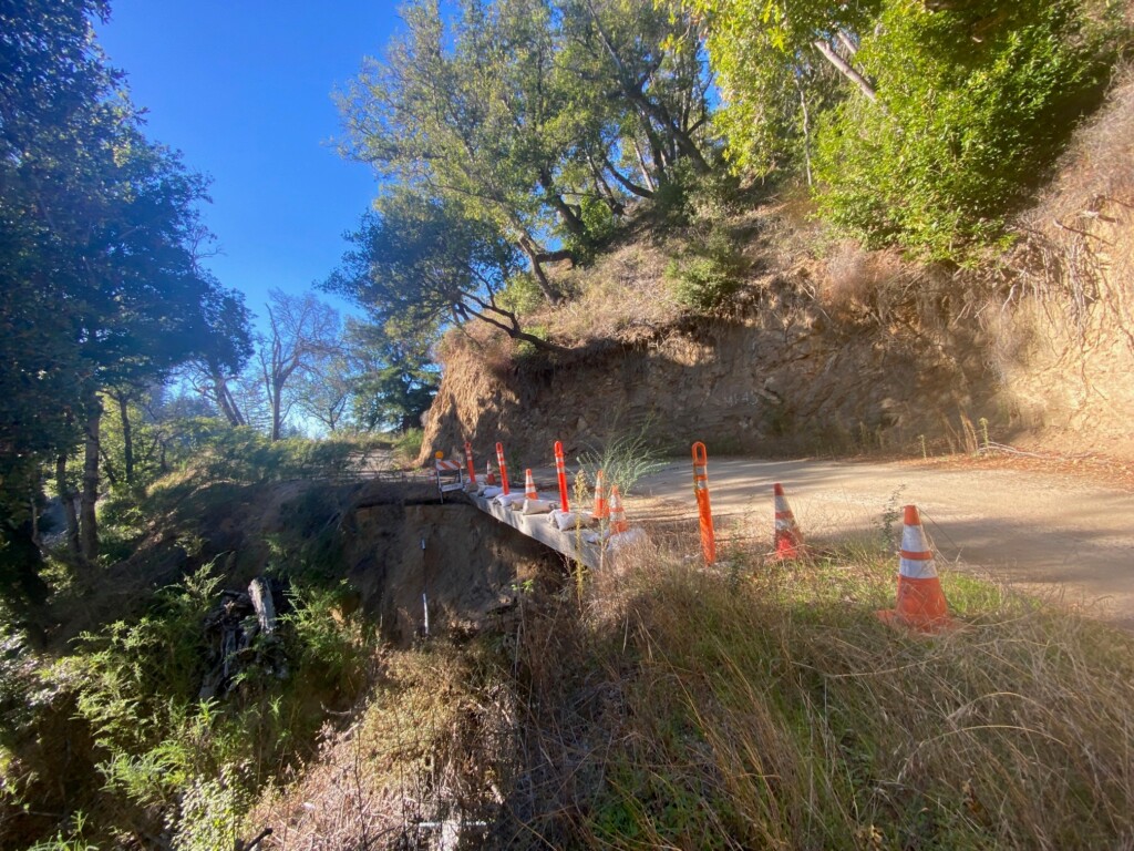 A washed-out road from storm damage, a road is seen halfway destroyed with cones aligning with the cracked area. 