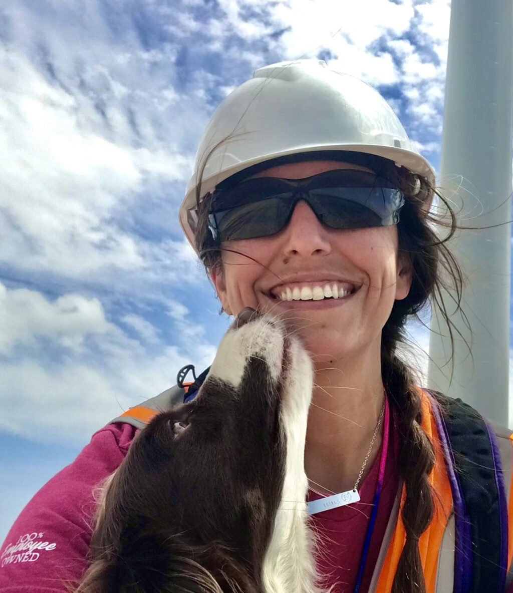 Smiling woman in hard hat and sunglasses gets nuzzled by a dog.