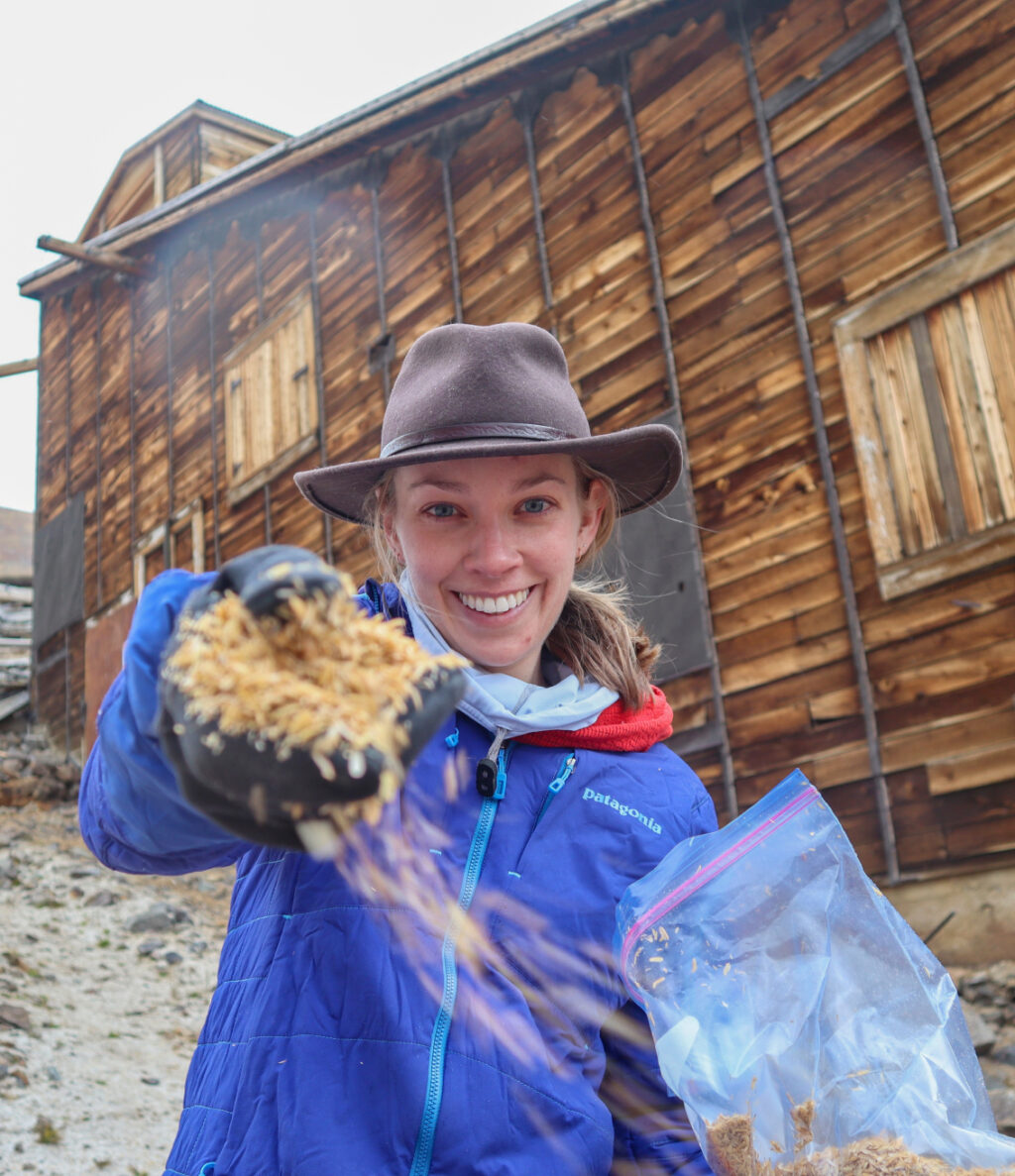 A smiling person holding feed for animals with a scooper and plastic bag, in front of an old wooden building.