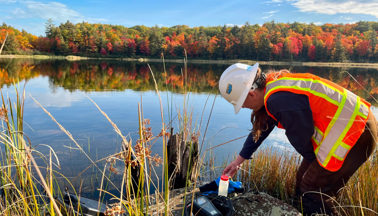 Worker in safety vest at lakeside, examining water collection kit.