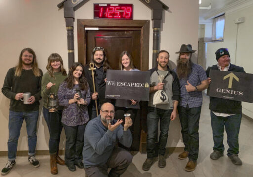 Nine people posing outside the escape room door, holding the sign "We Escaped".