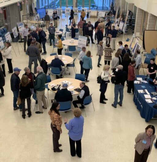 High-angle photo of an indoor public meeting. There are many people standing, posters around the room, and tables with papers on them.
