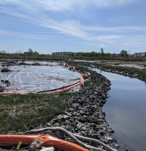 Small pond area affected by a spillage incident. Orange containment booms visible on water and land.