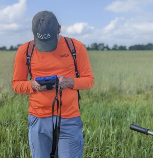A person in tall grass, wearing an orange SWCA jersey and cap, looks down at a digital device in his hands.