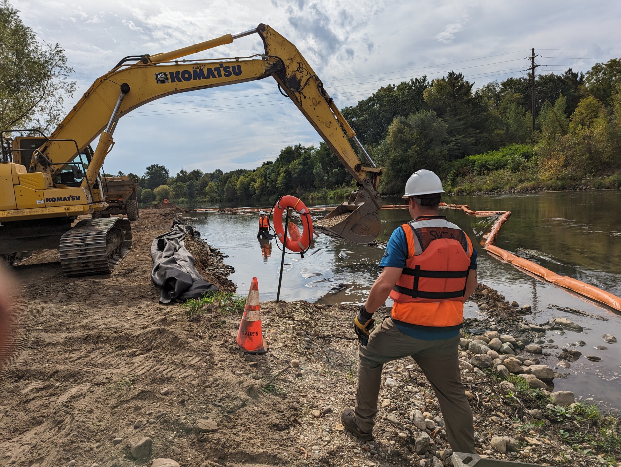 Staff member in an orange safety vest standing on the bank of a river in front of heavy construction equipment for a dam removal project.