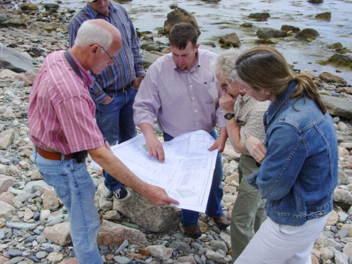 At a rocky shoreline, four people read from a diagram two of them are holding.