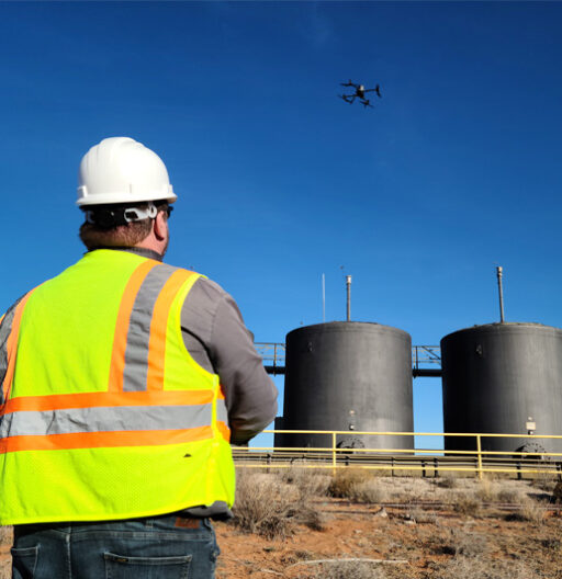 A man in a hard hat and safety vest is flying a drone over cylindrical tanks.