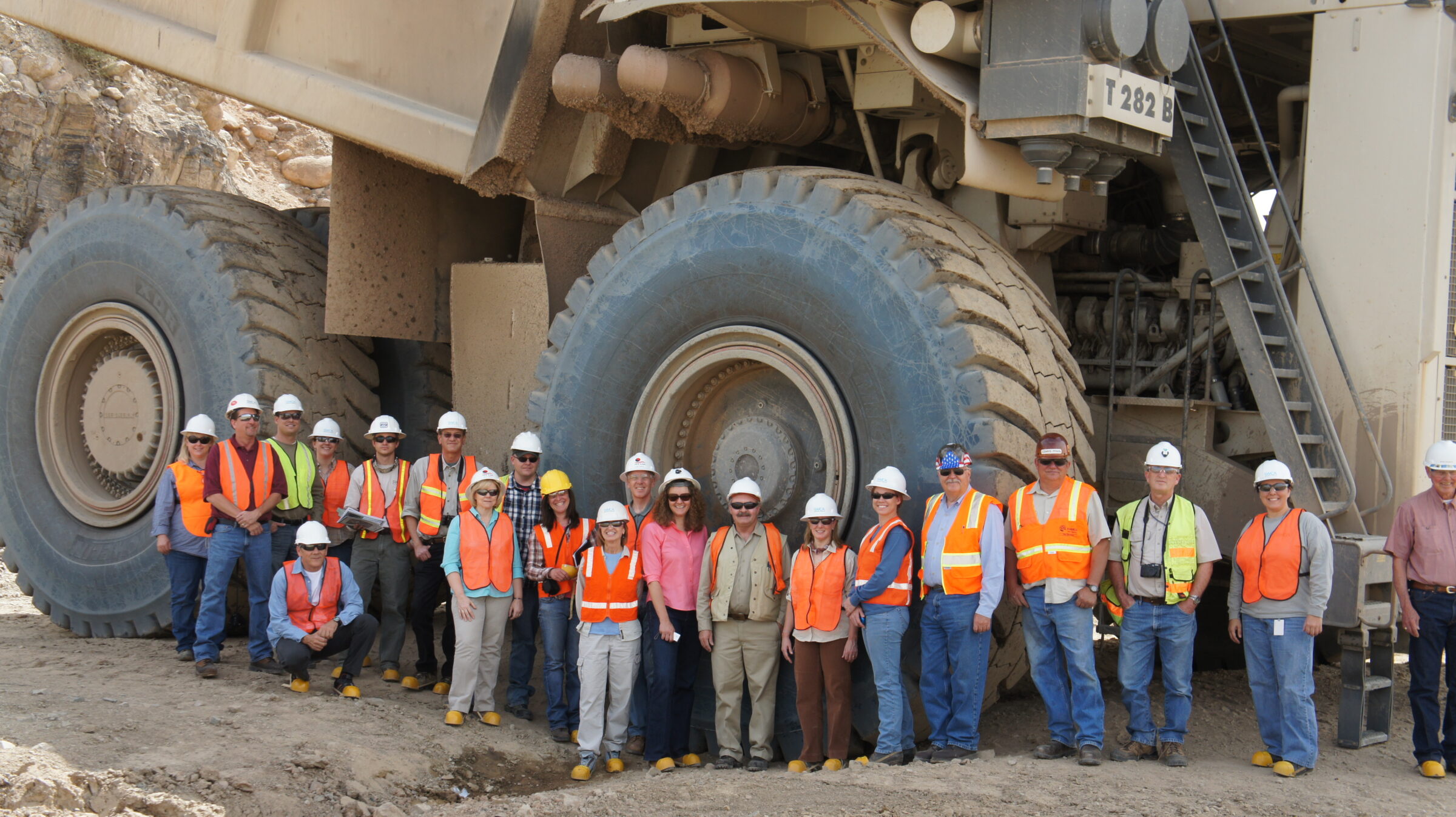 A group of over twenty people stand in front of a giant machine whose tire is twice their height.