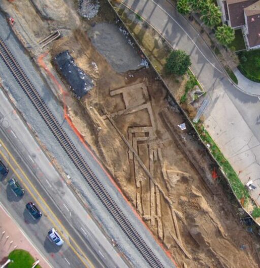 Aerial view of a long trench with exposed pipes, dug between a road and a sidewalk.