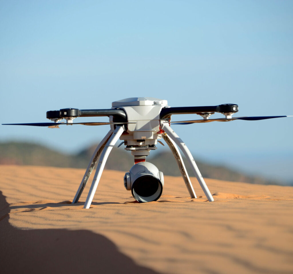 Close-up shot of a drone with four legs and four propellers, resting on the sand, and a bottom-mounted camera.