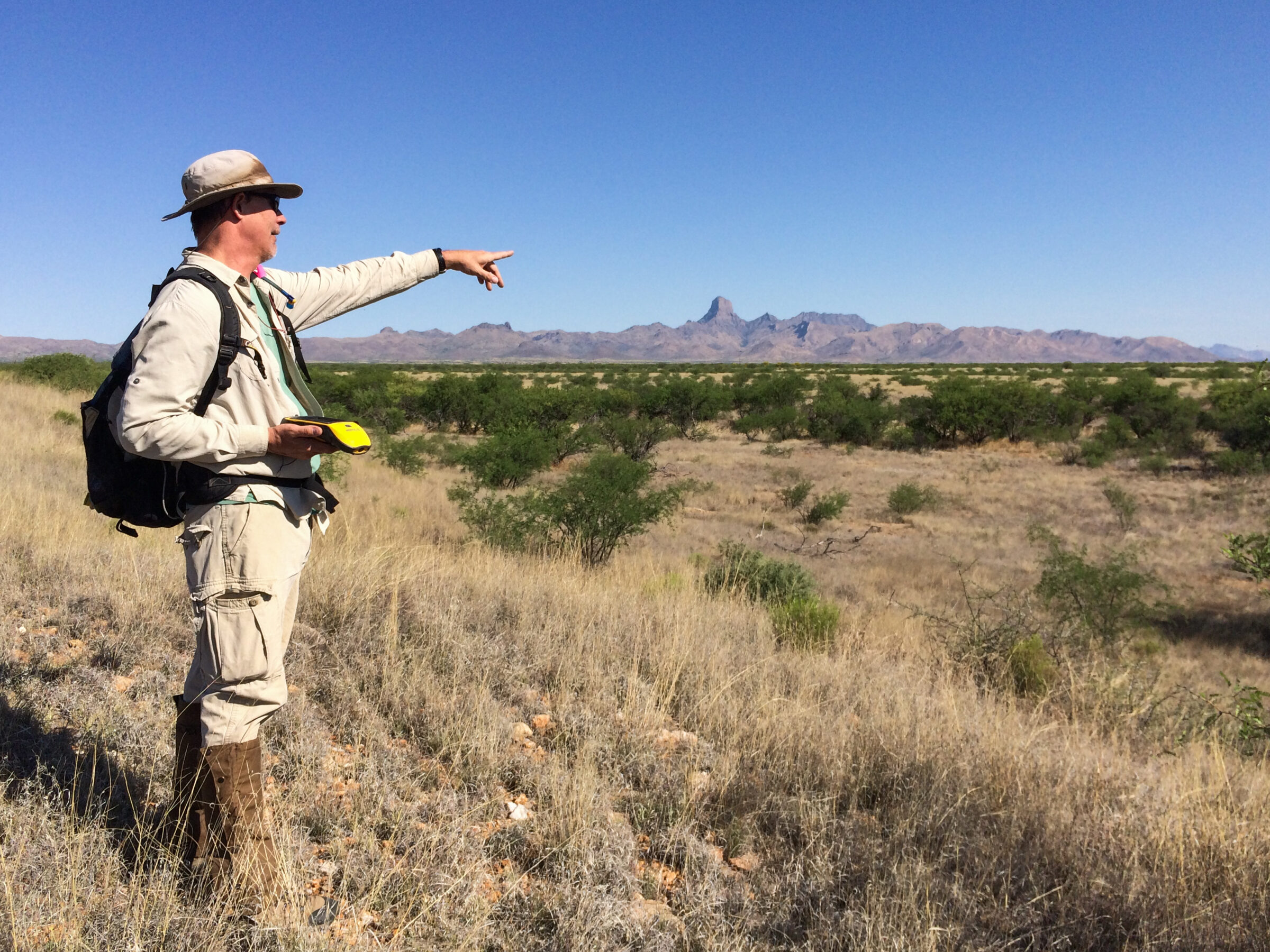 One worker in a field of dry grass points to a distant mountain formation.