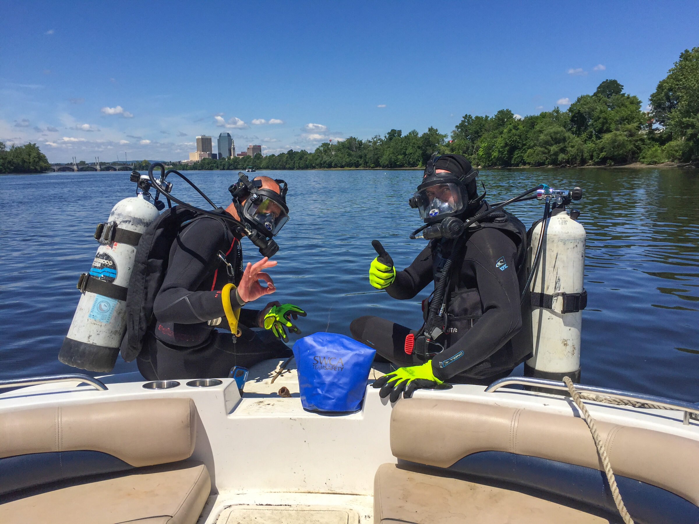 Two divers in full diving gear with oxygen tanks on their backs sit on the edge of a boat and wave.