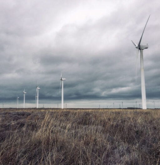 A ground-angle view of five wind turbines receding into the distance while storm-clouds form overhead.