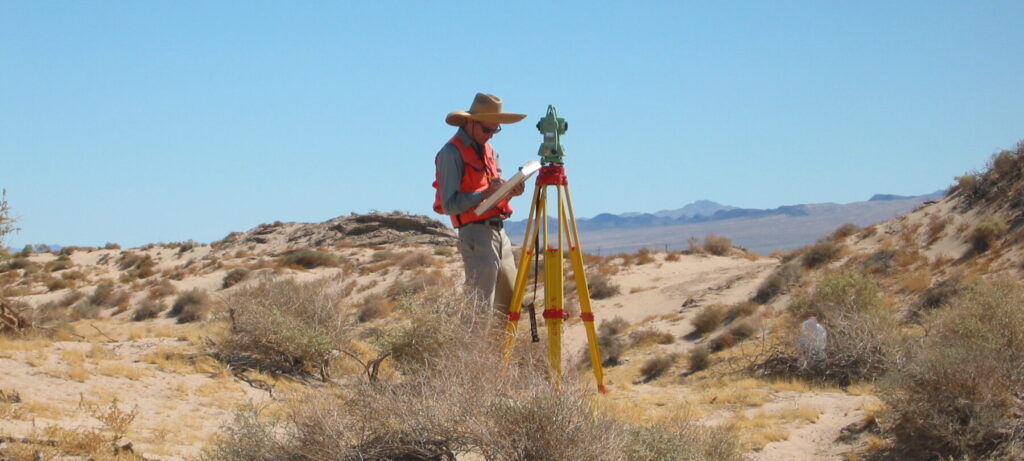 A man writes on a clipboard while standing next to a survey unit on a tall tripod, in an arid, dry landscape.